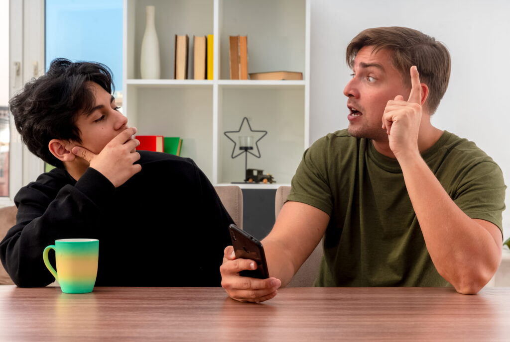 surprised young blonde handsome man points up holding phone sitting table looking each other with confident young brunette handsome guy putting hand chin inside living room 1