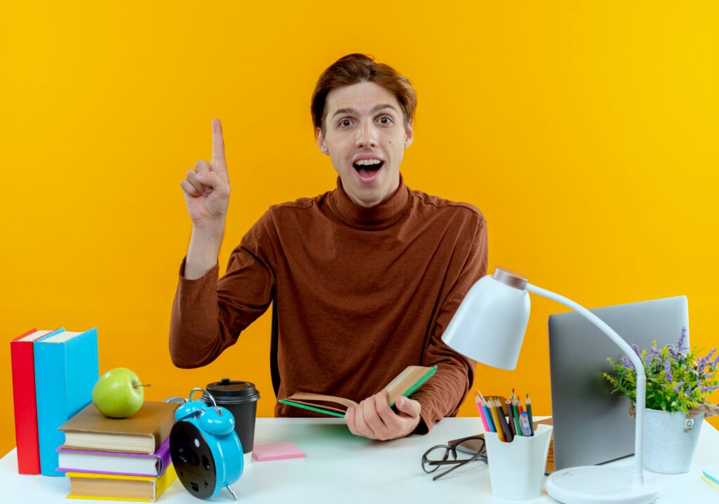 surprised young student boy sitting desk with school tools holding book points up yellow